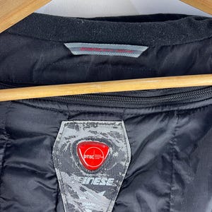 K&ouml;nnte beinhalten: Schwarz gesteppte Jacke mit schwarzem Kragen und rotem und silbernem Logoaufn&auml;her. Auf dem Aufn&auml;her steht das Wort "D-TEC" und der Markenname "Dainese". Die Jacke h&auml;ngt an einem Holzb&uuml;gel.