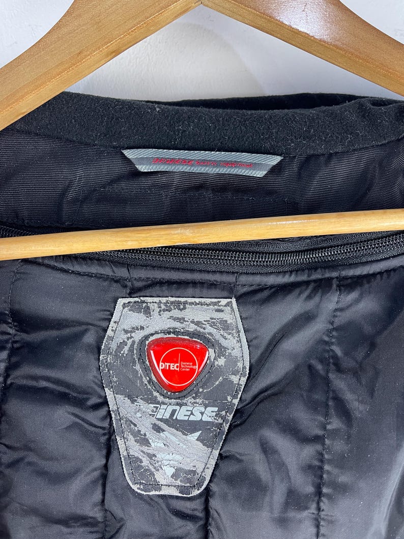 K&ouml;nnte beinhalten: Schwarz gesteppte Jacke mit schwarzem Kragen und rotem und silbernem Logoaufn&auml;her. Auf dem Aufn&auml;her steht das Wort "D-TEC" und der Markenname "Dainese". Die Jacke h&auml;ngt an einem Holzb&uuml;gel.