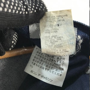 以下が含まれることがあります： 白い文字で書かれた青いデニム生地のラベルのクローズアップ。ラベルには、「COMME des GARCONS」、「MADE IN JAPAN」、「FABRIQUE AU JAPON」、「HP-01903M」、「100% COTTON」、「100% COTON」、「HAND WASH」、「DO NOT BLEACH」、「USE COOL IRON」、「DRY CLEAN」、「LINE DRY」、「LAVAGE MAINS」、「JAVEL INTERDIT」、「FER DOUX」、「NETTOYAGE A SEC」、「SECHAGE A LOMBRE」と記載されています。ラベルには日本語のテキストも含まれています。