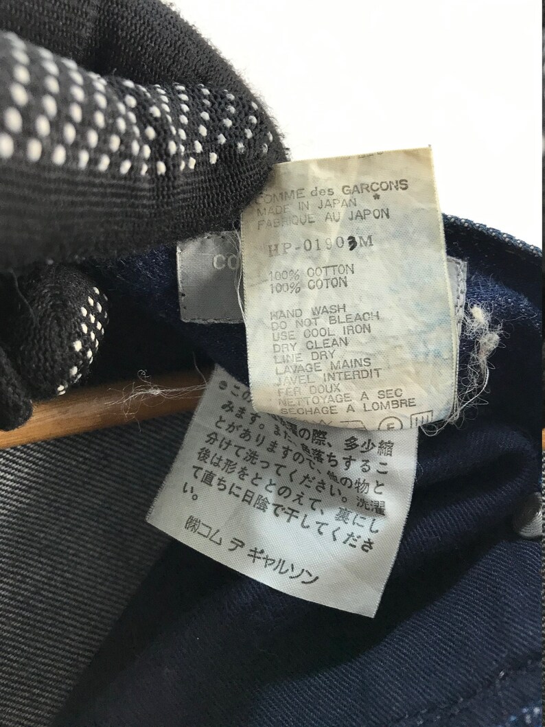 以下が含まれることがあります： 白い文字で書かれた青いデニム生地のラベルのクローズアップ。ラベルには、「COMME des GARCONS」、「MADE IN JAPAN」、「FABRIQUE AU JAPON」、「HP-01903M」、「100% COTTON」、「100% COTON」、「HAND WASH」、「DO NOT BLEACH」、「USE COOL IRON」、「DRY CLEAN」、「LINE DRY」、「LAVAGE MAINS」、「JAVEL INTERDIT」、「FER DOUX」、「NETTOYAGE A SEC」、「SECHAGE A LOMBRE」と記載されています。ラベルには日本語のテキストも含まれています。
