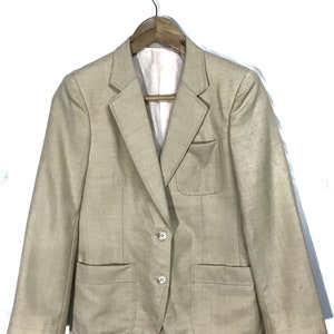 Puede incluir: Una chaqueta beige con dos botones de cierre y dos bolsillos de parche. La chaqueta está colgada en una percha de madera.