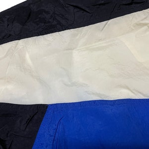 Op de afbeelding: Een zwart, wit en blauw nylon jack met een ritssluiting. Het jack heeft een kleurgeblokt ontwerp met een wit paneel in het midden.