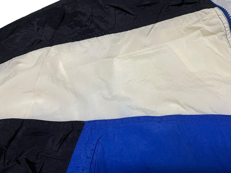 Op de afbeelding: Een zwart, wit en blauw nylon jack met een ritssluiting. Het jack heeft een kleurgeblokt ontwerp met een wit paneel in het midden.