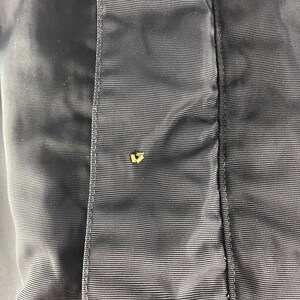 K&ouml;nnte beinhalten: Nahaufnahme eines Abschnitts einer dunkelblauen Jacke. Der Stoff hat einen subtilen Glanz und eine feine, horizontale Textur. Ein kleiner, goldfarbener Knopf ist sichtbar und setzt einen Kontrast zu dem ansonsten monochromen Design.