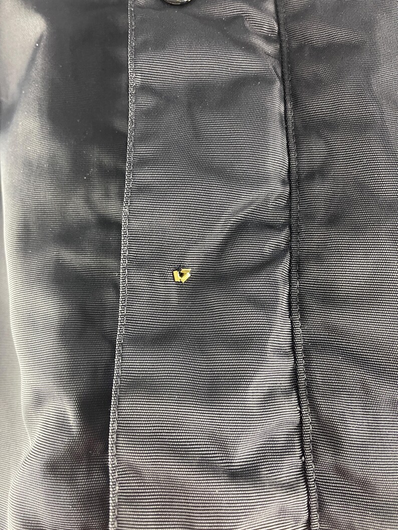 K&ouml;nnte beinhalten: Nahaufnahme eines Abschnitts einer dunkelblauen Jacke. Der Stoff hat einen subtilen Glanz und eine feine, horizontale Textur. Ein kleiner, goldfarbener Knopf ist sichtbar und setzt einen Kontrast zu dem ansonsten monochromen Design.