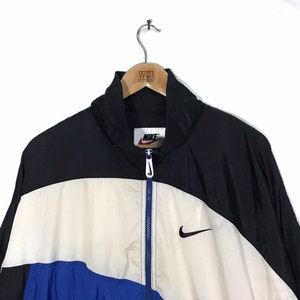 Op de afbeelding: Een zwart, wit en blauw windjack met een ritssluiting. Het jack heeft een Nike-logo op de linkerborst en een Nike-swoosh aan de rechterkant.