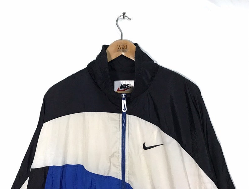 Op de afbeelding: Een zwart, wit en blauw windjack met een ritssluiting. Het jack heeft een Nike-logo op de linkerborst en een Nike-swoosh aan de rechterkant.