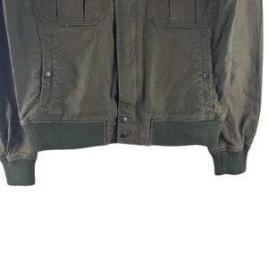 May include: Olive green bomber jacket with ribbed cuffs and hem. Features two front pockets with button closures and two chest pockets. The jacket has a button-up front and a classic bomber style.