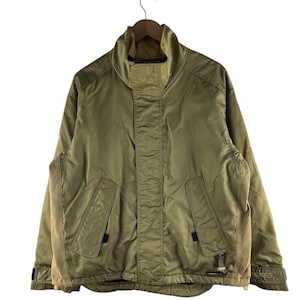 May include: A tan jacket with a high collar and two front pockets. The jacket has a zipper closure and a contrasting fabric panel on the sides. The jacket is hanging on a wooden hanger against a white background.