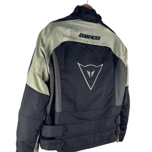 K&ouml;nnte beinhalten: Schwarze und graue Motorradjacke mit dem Markennamen "DAINESSE" auf dem R&uuml;cken aufgedruckt. Die Jacke hat ein wei&szlig;es Logo auf dem R&uuml;cken und verstellbare Taillenriemen. Die Jacke h&auml;ngt an einem Metallb&uuml;gel.