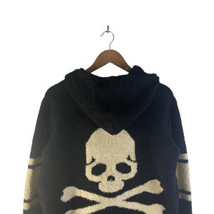 May include: Black hooded sweater with a large skull and crossbones design on the back. The skull is off-white, and the sweater has tan stripes on the sleeves.