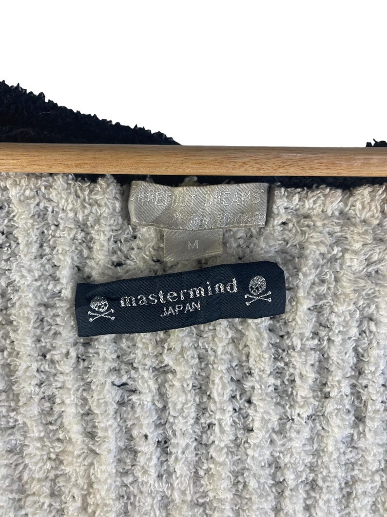 May include: Close-up of a textured, off-white and black fabric. The image shows two tags: one with the brand "Barefoot Dreams" and size "M", and another black tag with the text "mastermind JAPAN" and a skull and crossbones logo.