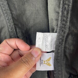May include: Close-up of a dark green jacket with a tag held by a hand. The tag displays the Avirex logo, a gold emblem with wings, and the words "AUTHENTIC PRODUCT". The jacket's fabric is visible, showing a textured weave.