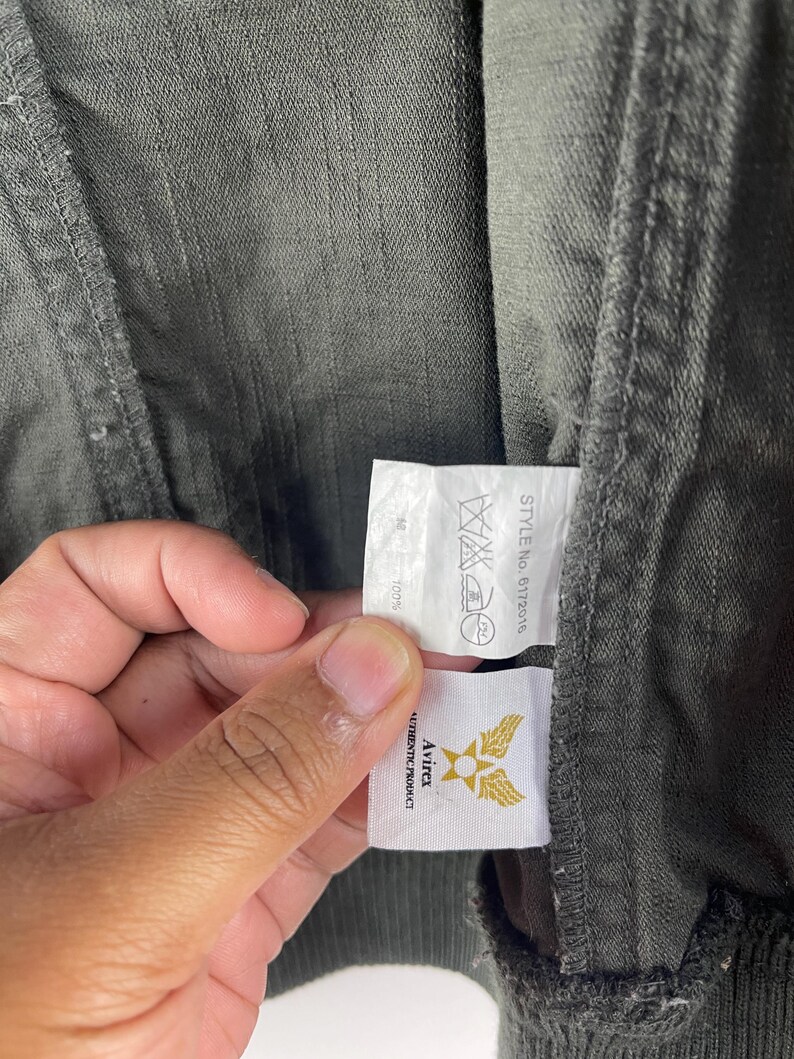 May include: Close-up of a dark green jacket with a tag held by a hand. The tag displays the Avirex logo, a gold emblem with wings, and the words "AUTHENTIC PRODUCT". The jacket's fabric is visible, showing a textured weave.