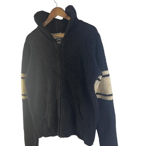 May include: Black zip-up hoodie with a textured, fuzzy appearance. The sleeves feature beige stripes. The hoodie has a hood and two front pockets. The jacket is hanging on a wooden hanger against a white background.