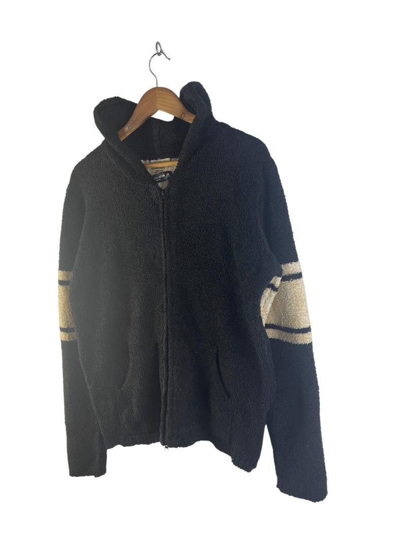 May include: Black zip-up hoodie with a textured, fuzzy appearance. The sleeves feature beige stripes. The hoodie has a hood and two front pockets. The jacket is hanging on a wooden hanger against a white background.