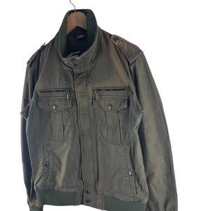 May include: Olive green bomber jacket with a stand-up collar, multiple front pockets, and a ribbed hem and cuffs. The jacket is hanging on a wooden hanger against a white background. Features a zipper closure and button details.