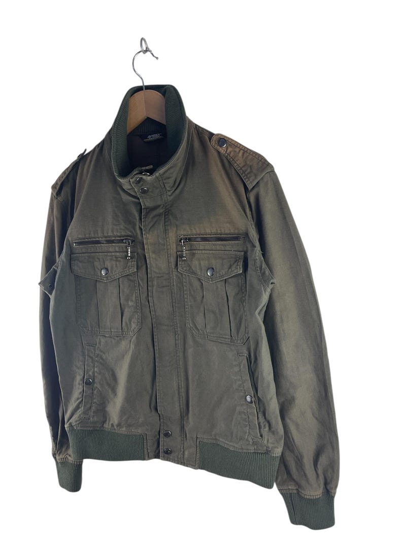 May include: Olive green bomber jacket with a stand-up collar, multiple front pockets, and a ribbed hem and cuffs. The jacket is hanging on a wooden hanger against a white background. Features a zipper closure and button details.