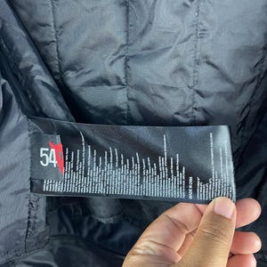 K&ouml;nnte beinhalten: Schwarze Jacke mit gestepptem Futter. Ein Etikett mit der Nummer "54" und Text in mehreren Sprachen ist sichtbar. Die Jacke scheint aus Nylon und Polyester zu bestehen.