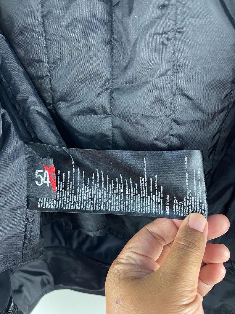 K&ouml;nnte beinhalten: Schwarze Jacke mit gestepptem Futter. Ein Etikett mit der Nummer "54" und Text in mehreren Sprachen ist sichtbar. Die Jacke scheint aus Nylon und Polyester zu bestehen.