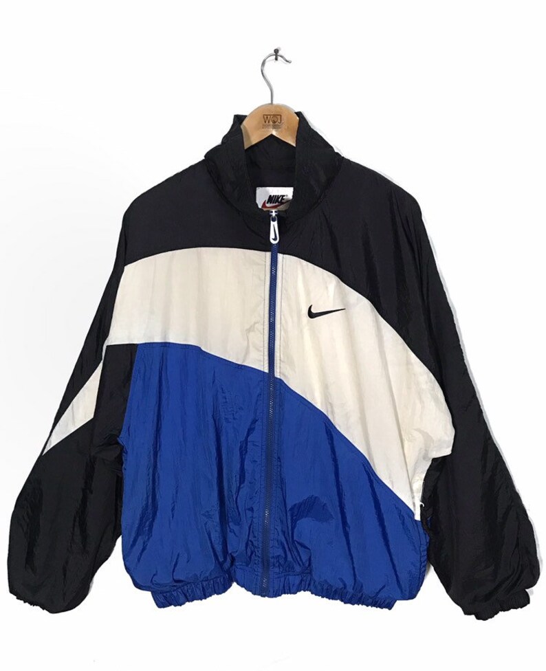 Op de afbeelding: Een zwart, wit en blauw windjack met een ritssluiting en een Nike-logo op de rechterborst.