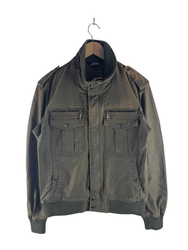May include: Olive green bomber jacket with a high collar and multiple pockets. The jacket features a ribbed hem and cuffs, and a zipper closure. The jacket is hanging on a wooden hanger against a white background.