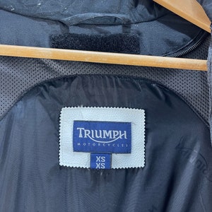 Puede incluir: Chaqueta de moto negra con un parche de cuero blanco que dice "Triumph Motorcycles" y una etiqueta de talla que dice "XS".