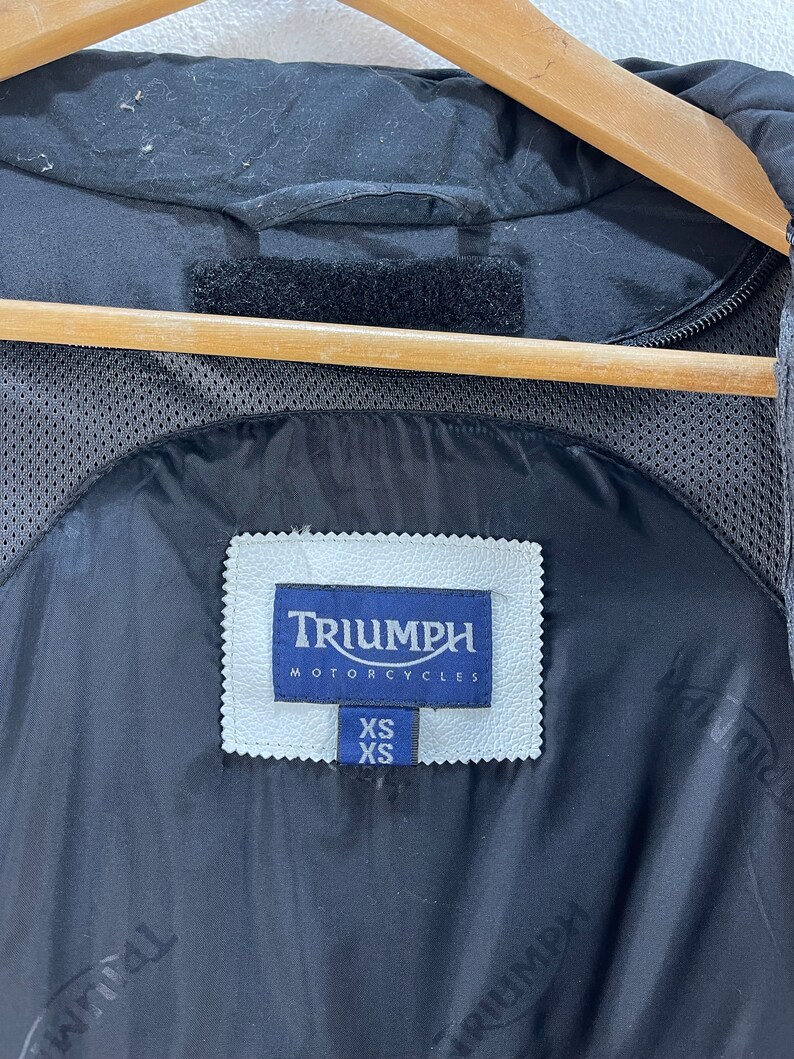 Puede incluir: Chaqueta de moto negra con un parche de cuero blanco que dice "Triumph Motorcycles" y una etiqueta de talla que dice "XS".