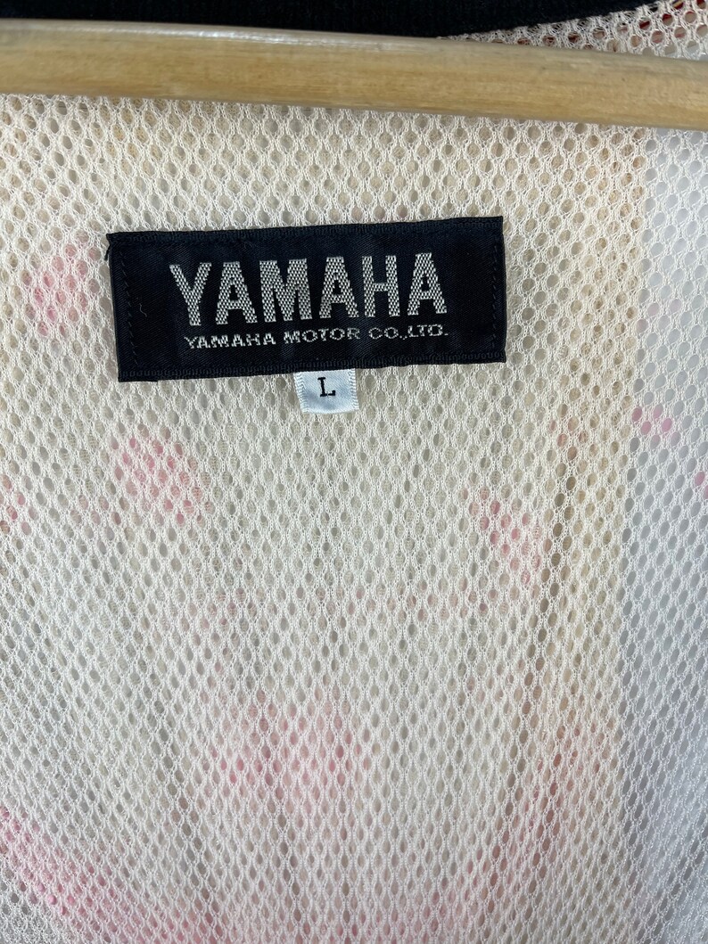 Puede incluir: Una etiqueta en blanco y negro con el texto "YAMAHA" y "YAMAHA MOTOR CO., LTD." en blanco. La etiqueta tambi&eacute;n tiene una etiqueta de tama&ntilde;o "L" blanca.