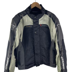 K&ouml;nnte beinhalten: Schwarze, graue und beige Motorradjacke mit dem Markennamen "Dainese" auf den &Auml;rmeln. Die Jacke hat einen Stehkragen, Druckkn&ouml;pfe und ein rotes Logo auf der Brust. Die Jacke h&auml;ngt an einem Holzb&uuml;gel.
