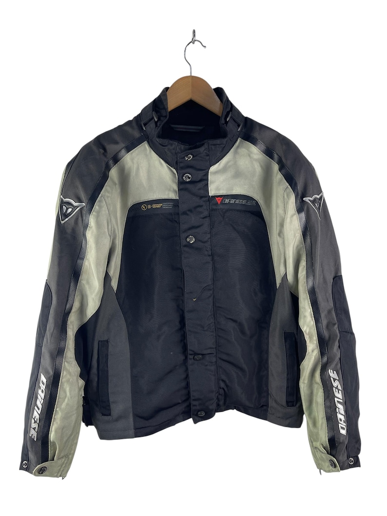 K&ouml;nnte beinhalten: Schwarze, graue und beige Motorradjacke mit dem Markennamen "Dainese" auf den &Auml;rmeln. Die Jacke hat einen Stehkragen, Druckkn&ouml;pfe und ein rotes Logo auf der Brust. Die Jacke h&auml;ngt an einem Holzb&uuml;gel.