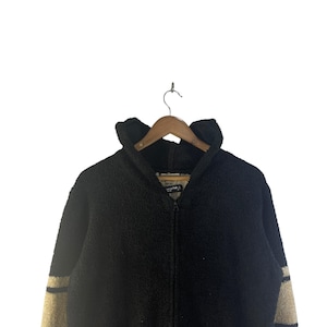 May include: A black zip-up jacket with a high collar, hanging on a wooden hanger. The jacket has beige stripes on the sleeves. The material appears to be a thick, textured fabric, suitable for cold weather.