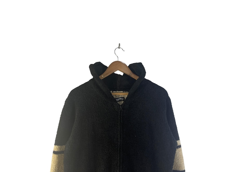 May include: A black zip-up jacket with a high collar, hanging on a wooden hanger. The jacket has beige stripes on the sleeves. The material appears to be a thick, textured fabric, suitable for cold weather.