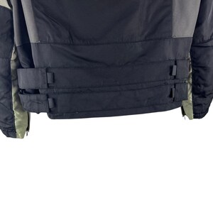 K&ouml;nnte beinhalten: Eine schwarze und graue Winterjacke mit hohem Kragen und Rei&szlig;verschluss. Die Jacke hat zwei schwarze Riemen mit Schnallen am unteren R&uuml;cken. Die &Auml;rmel sind schwarz und grau mit gr&uuml;nem Futter.