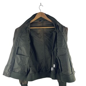 May include: Olive green bomber jacket with a ribbed collar and cuffs. The jacket has multiple pockets and a zipper closure. The interior lining is a darker shade of green. The jacket is hanging on a wooden hanger.