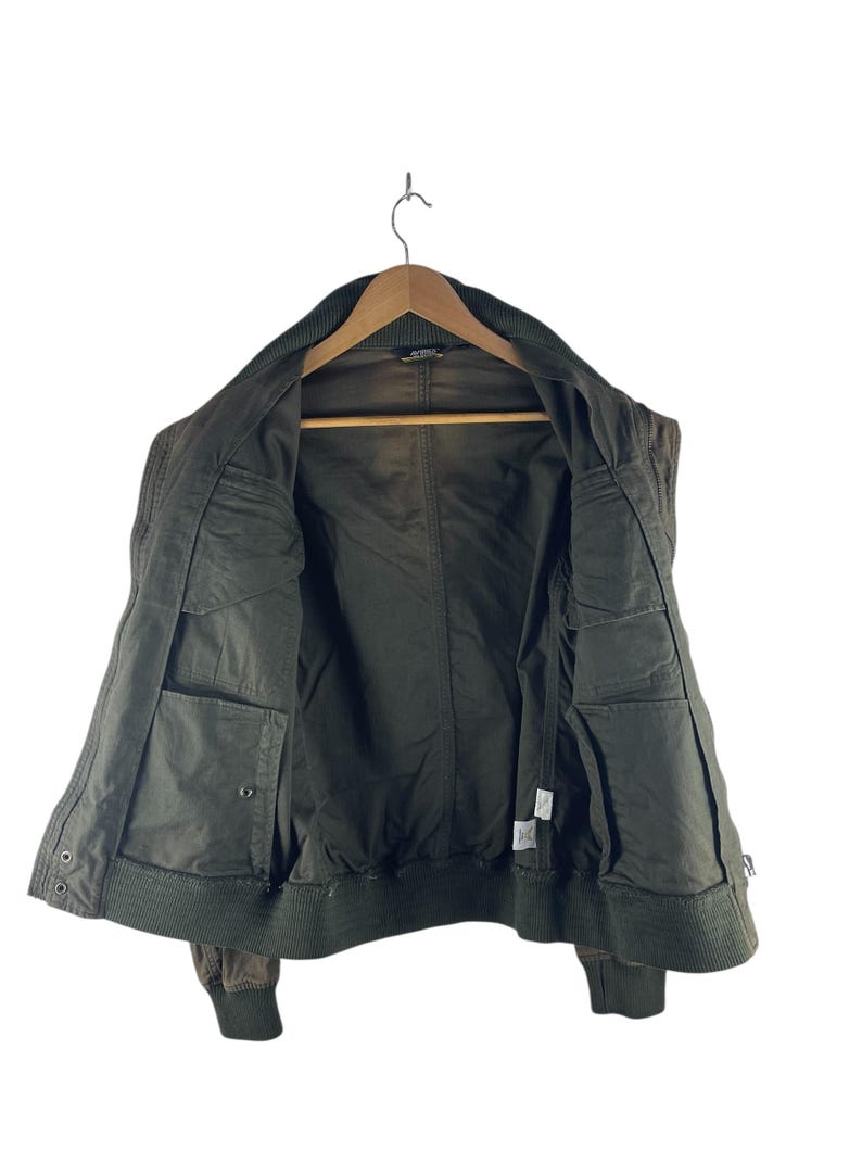 May include: Olive green bomber jacket with a ribbed collar and cuffs. The jacket has multiple pockets and a zipper closure. The interior lining is a darker shade of green. The jacket is hanging on a wooden hanger.