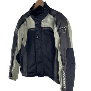 K&ouml;nnte beinhalten: Schwarze und graue Motorradjacke mit dem Markennamen "DAINESE" auf den &Auml;rmeln. Die Jacke hat einen Stehkragen, mehrere Taschen und ein rotes Logo auf der Brust. Sie h&auml;ngt an einem Holzkleiderb&uuml;gel.