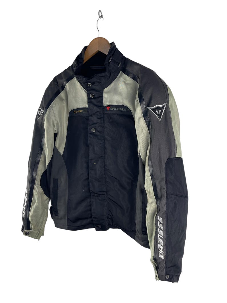 K&ouml;nnte beinhalten: Schwarze und graue Motorradjacke mit dem Markennamen "DAINESE" auf den &Auml;rmeln. Die Jacke hat einen Stehkragen, mehrere Taschen und ein rotes Logo auf der Brust. Sie h&auml;ngt an einem Holzkleiderb&uuml;gel.