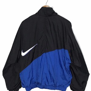 Op de afbeelding: Een zwart en blauw windjack met een wit Nike swoosh-logo aan de linkerkant.