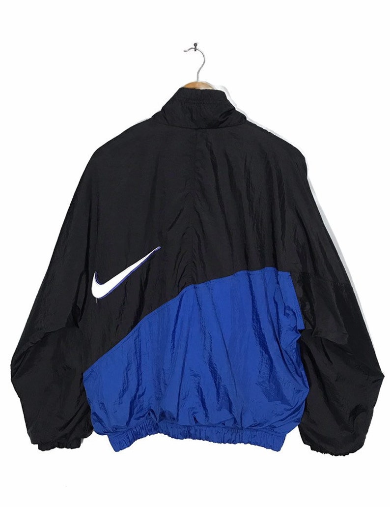 Op de afbeelding: Een zwart en blauw windjack met een wit Nike swoosh-logo aan de linkerkant.