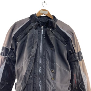 Puede incluir: Una chaqueta de moto negra y gris con cierre de cremallera y m&uacute;ltiples bolsillos. La chaqueta tiene una banda reflectante en los hombros y los brazos.