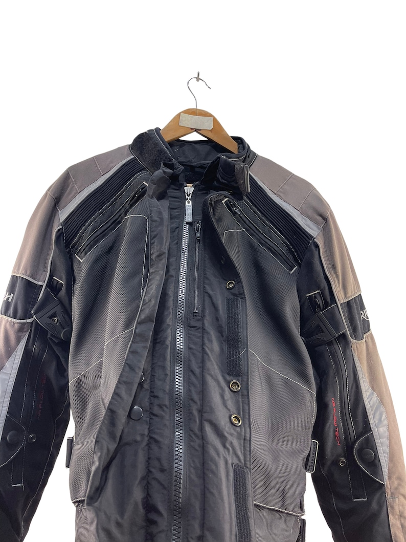 Puede incluir: Una chaqueta de moto negra y gris con cierre de cremallera y m&uacute;ltiples bolsillos. La chaqueta tiene una banda reflectante en los hombros y los brazos.
