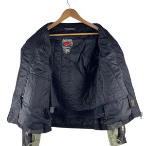 K&ouml;nnte beinhalten: Schwarze Motorradjacke mit gestepptem Innenfutter, h&auml;ngt an einem Holzb&uuml;gel. Die Jacke hat mehrere Taschen, einen Rei&szlig;verschluss und Akzente in Grau und Olivgr&uuml;n an den Manschetten. Ein Logo ist auf der Brust sichtbar.