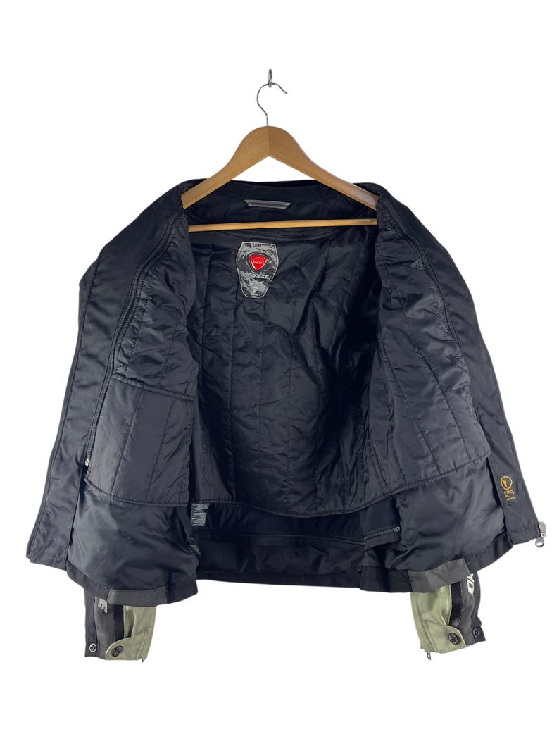 K&ouml;nnte beinhalten: Schwarze Motorradjacke mit gestepptem Innenfutter, h&auml;ngt an einem Holzb&uuml;gel. Die Jacke hat mehrere Taschen, einen Rei&szlig;verschluss und Akzente in Grau und Olivgr&uuml;n an den Manschetten. Ein Logo ist auf der Brust sichtbar.