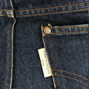 以下が含まれることがあります： 濃い青色のデニムのポケットのクローズアップ。白いラベルには「COMME des GARCONS HOMME*」と書いてあります。