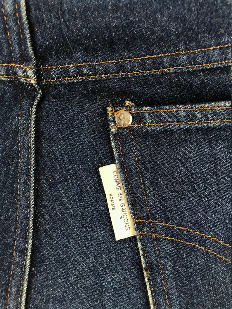 以下が含まれることがあります： 濃い青色のデニムのポケットのクローズアップ。白いラベルには「COMME des GARCONS HOMME*」と書いてあります。