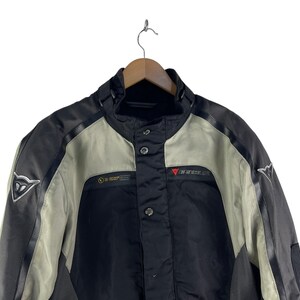 K&ouml;nnte beinhalten: Schwarze und graue Motorradjacke mit hohem Kragen und Knopfverschluss. Die Jacke hat ein D-air-Logo auf der Schulter und das Dainese-Logo auf der Brust. Die Jacke h&auml;ngt an einem Holzb&uuml;gel.