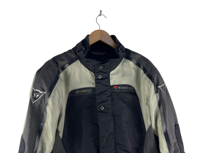 K&ouml;nnte beinhalten: Schwarze und graue Motorradjacke mit hohem Kragen und Knopfverschluss. Die Jacke hat ein D-air-Logo auf der Schulter und das Dainese-Logo auf der Brust. Die Jacke h&auml;ngt an einem Holzb&uuml;gel.