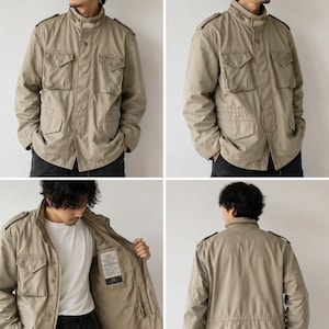 May include: A beige field jacket featuring a stand-up collar, epaulettes on the shoulders, and four front flap pockets. The jacket includes a front zip closure with a snap-button placket, long sleeves, and an interior label with visible text.