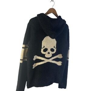 May include: Black hooded sweater with a skull and crossbones design on the back. The sweater has gold stripes on the sleeves and a matching gold skull graphic. The sweater is hanging on a wooden hanger.